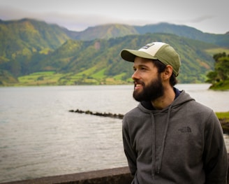 man in gray hoodie wearing white cap standing near body of water during daytime