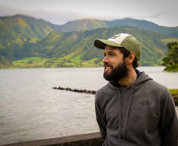 man in gray hoodie wearing white cap standing near body of water during daytime