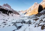 A snowy alpine landscape with rustic stone buildings nestled in a valley. The surrounding mountains have a mix of snow and exposed rock, with tall, dark evergreen trees scattered along the slopes. The sky is clear and blue, adding a serene and tranquil atmosphere to the scene.
