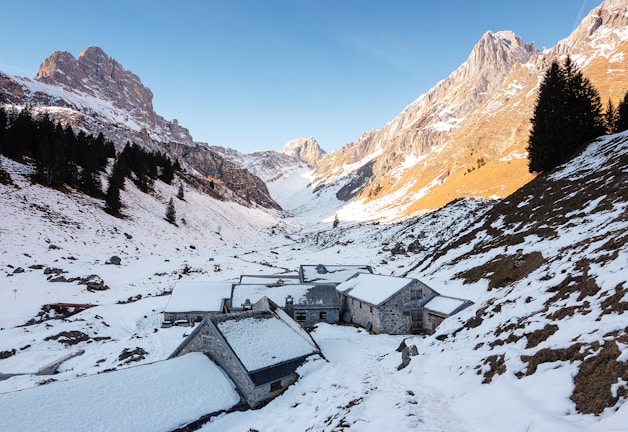 A snowy alpine landscape with rustic stone buildings nestled in a valley. The surrounding mountains have a mix of snow and exposed rock, with tall, dark evergreen trees scattered along the slopes. The sky is clear and blue, adding a serene and tranquil atmosphere to the scene.
