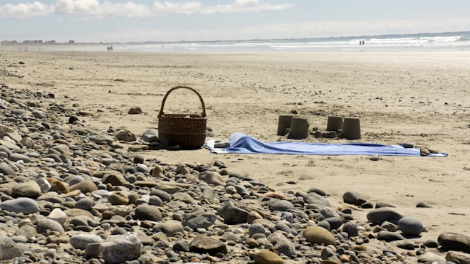 A sandy beach with scattered pebbles in the foreground, featuring a blue towel and a wicker picnic basket. Several sandcastles are built nearby, and the distant shoreline is visible with gentle waves. People can be seen walking along the water's edge under a partly cloudy sky.