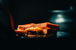 A grilled cheese sandwich is positioned on a dark surface, with light softly illuminating the crispy golden-brown crust. Melted cheese oozes slightly between the slices, creating a warm and inviting texture. The background is softly blurred, emphasizing the sandwich.