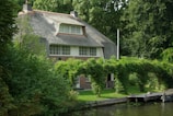 A charming cottage with a thatched roof surrounded by lush greenery. It features large windows and is situated next to a waterway. The garden is vibrant with well-maintained plants and an arched trellis covered with foliage.