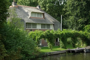 A charming cottage with a thatched roof surrounded by lush greenery. It features large windows and is situated next to a waterway. The garden is vibrant with well-maintained plants and an arched trellis covered with foliage.