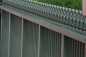 A close-up view of a metal fence with vertical bars and a decorative top section featuring a repeating pattern. The fence has a greenish hue and a slightly weathered texture.