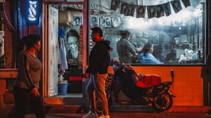 A nighttime street scene depicting a small, illuminated hair salon. Inside the glass storefront, a hairdresser is attending to a client while various photographs of hairstyles are displayed along the wall. Outside, a man and a woman walk past, and a motorbike is parked in front with a red cover.