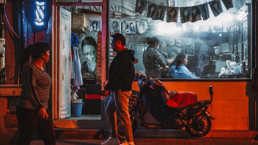 A nighttime street scene depicting a small, illuminated hair salon. Inside the glass storefront, a hairdresser is attending to a client while various photographs of hairstyles are displayed along the wall. Outside, a man and a woman walk past, and a motorbike is parked in front with a red cover.