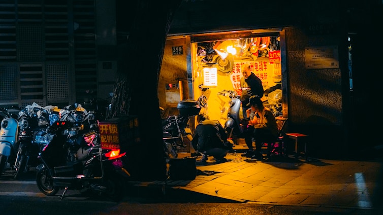 A small, illuminated repair shop is nestled within a dimly lit urban environment. The shop is busy with activity as a person repairs a scooter and another individual sits nearby, seemingly engaged in conversation or rest. Various scooters are parked outside, some with visible signage, while the shop's interior is cluttered with tools and parts.