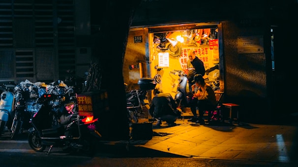 A small, illuminated repair shop is nestled within a dimly lit urban environment. The shop is busy with activity as a person repairs a scooter and another individual sits nearby, seemingly engaged in conversation or rest. Various scooters are parked outside, some with visible signage, while the shop's interior is cluttered with tools and parts.