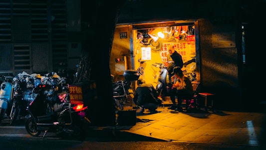 A small, illuminated repair shop is nestled within a dimly lit urban environment. The shop is busy with activity as a person repairs a scooter and another individual sits nearby, seemingly engaged in conversation or rest. Various scooters are parked outside, some with visible signage, while the shop's interior is cluttered with tools and parts.