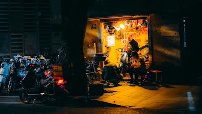 A small, illuminated repair shop is nestled within a dimly lit urban environment. The shop is busy with activity as a person repairs a scooter and another individual sits nearby, seemingly engaged in conversation or rest. Various scooters are parked outside, some with visible signage, while the shop's interior is cluttered with tools and parts.
