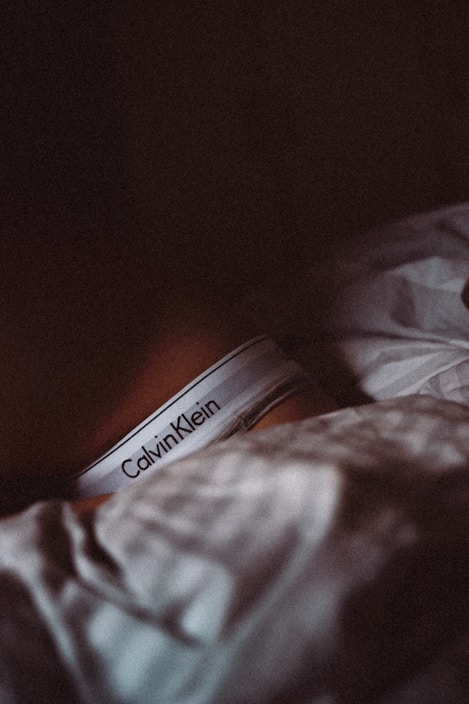 A close-up view of a person partially covered by a blanket, wearing Calvin Klein underwear in a dimly lit setting.