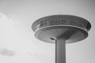 A futuristic, elevated structure with a round, disc-shaped observation deck supported by a central column. The sleek design is accentuated by horizontal lines and metallic surfaces.