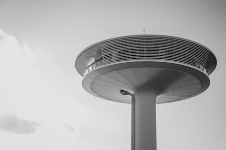 A futuristic, elevated structure with a round, disc-shaped observation deck supported by a central column. The sleek design is accentuated by horizontal lines and metallic surfaces.