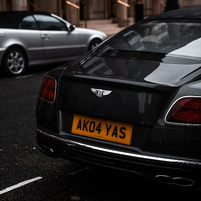 A dark-colored luxury car is parked on a street with a visible license plate reading 'AK04 YAS'. The car displays the Bentley logo prominently on its back. In the background, there is another silver-colored car partially visible. The setting appears to be urban, with building structures and steps in the background.