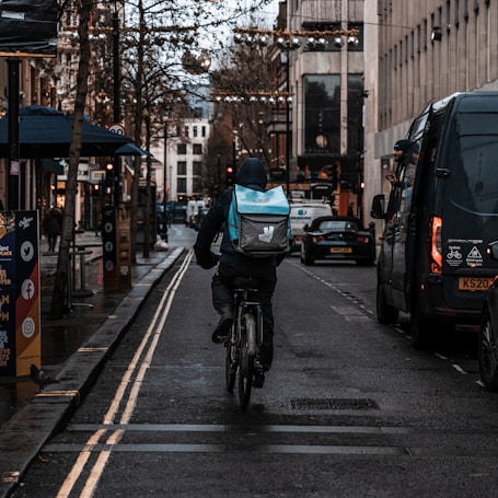 A person on a bicycle rides down a city street with a large backpack, likely used for deliveries. The street is lined with parked vehicles and shops, and the atmosphere seems damp and overcast. Decorative lights hang across the street, and there are trees lining the sidewalk.