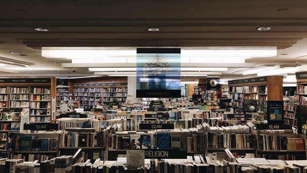 A large bookstore with numerous bookshelves organized by categories such as humanities, religion, new age, and law. The shelves are densely packed with a wide variety of books. Overhead lights illuminate the space, creating a cozy and inviting atmosphere.