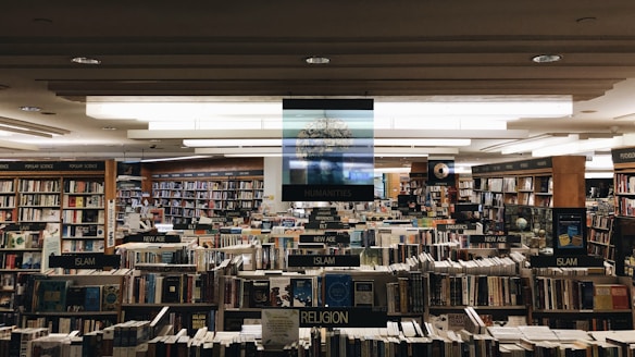 A large bookstore with numerous bookshelves organized by categories such as humanities, religion, new age, and law. The shelves are densely packed with a wide variety of books. Overhead lights illuminate the space, creating a cozy and inviting atmosphere.