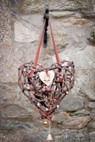 A rustic heart-shaped wreath made of interwoven sticks and decorated with small wooden stars, a wooden heart with a Christmas tree cutout, two purple spherical ornaments, and a small gold bauble. The wreath is hung on a metal hook against a textured stone wall with a reddish-brown ribbon.