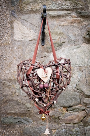 A rustic heart-shaped wreath made of interwoven sticks and decorated with small wooden stars, a wooden heart with a Christmas tree cutout, two purple spherical ornaments, and a small gold bauble. The wreath is hung on a metal hook against a textured stone wall with a reddish-brown ribbon.