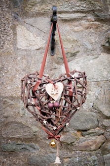 A rustic heart-shaped wreath made of interwoven sticks and decorated with small wooden stars, a wooden heart with a Christmas tree cutout, two purple spherical ornaments, and a small gold bauble. The wreath is hung on a metal hook against a textured stone wall with a reddish-brown ribbon.