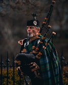A man dressed in traditional Scottish attire plays a bagpipe. He wears a dark uniform with intricate gold details and has a green and blue tartan draped across his shoulder. The bagpipe is ornate, made of dark leather with bright fittings. In the background, there is an iron fence and blurred foliage.