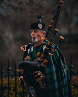 A man dressed in traditional Scottish attire plays a bagpipe. He wears a dark uniform with intricate gold details and has a green and blue tartan draped across his shoulder. The bagpipe is ornate, made of dark leather with bright fittings. In the background, there is an iron fence and blurred foliage.