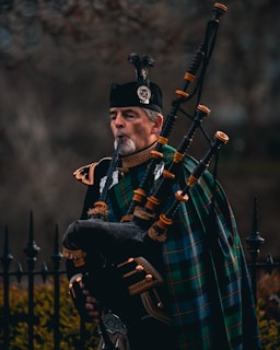 A man dressed in traditional Scottish attire plays a bagpipe. He wears a dark uniform with intricate gold details and has a green and blue tartan draped across his shoulder. The bagpipe is ornate, made of dark leather with bright fittings. In the background, there is an iron fence and blurred foliage.