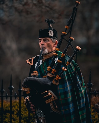 A man dressed in traditional Scottish attire plays a bagpipe. He wears a dark uniform with intricate gold details and has a green and blue tartan draped across his shoulder. The bagpipe is ornate, made of dark leather with bright fittings. In the background, there is an iron fence and blurred foliage.