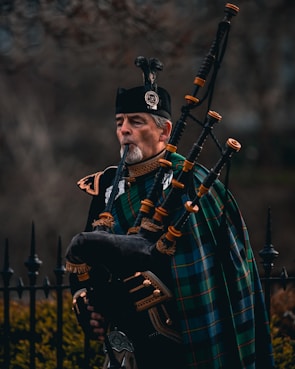 A man dressed in traditional Scottish attire plays a bagpipe. He wears a dark uniform with intricate gold details and has a green and blue tartan draped across his shoulder. The bagpipe is ornate, made of dark leather with bright fittings. In the background, there is an iron fence and blurred foliage.