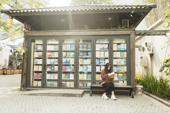A small wood-framed structure resembles a miniature library with glass doors, showcasing numerous books neatly arranged on its shelves. Outside, a person is seated on a bench reading a book. The setting is bright and outdoors, with plants and trees visible to the side.