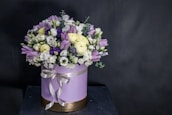 A round flower arrangement is placed in a cylindrical box decorated with a ribbon. The bouquet consists of various flowers including purple tulips, white roses, small daisies, and some deep violet blooms. The box is light purple with a golden base and a satin ribbon tied around it.