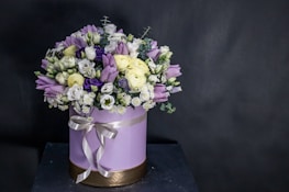 A round flower arrangement is placed in a cylindrical box decorated with a ribbon. The bouquet consists of various flowers including purple tulips, white roses, small daisies, and some deep violet blooms. The box is light purple with a golden base and a satin ribbon tied around it.