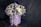 A round flower arrangement is placed in a cylindrical box decorated with a ribbon. The bouquet consists of various flowers including purple tulips, white roses, small daisies, and some deep violet blooms. The box is light purple with a golden base and a satin ribbon tied around it.