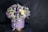 A round flower arrangement is placed in a cylindrical box decorated with a ribbon. The bouquet consists of various flowers including purple tulips, white roses, small daisies, and some deep violet blooms. The box is light purple with a golden base and a satin ribbon tied around it.