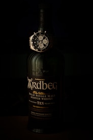A dark bottle of Ardbeg Islay Single Malt Scotch Whisky is prominently displayed, featuring detailed labeling that highlights it as ten years old. Positioned horizontally across the neck of the bottle is a sophisticated wristwatch with a black dial and chronograph features, adding a luxurious touch to the composition.