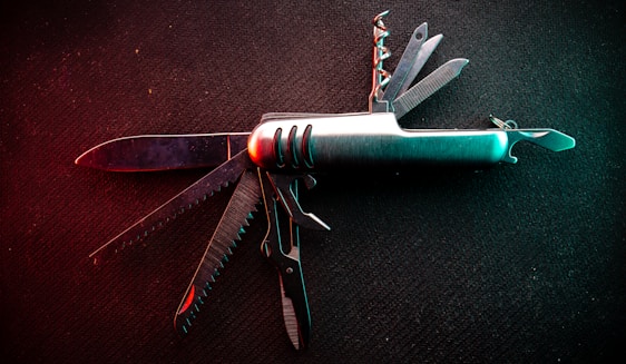 A multi-tool pocket knife is displayed with its various tools extended against a dark background. The knife includes multiple blades, a corkscrew, a saw, and other utility tools. The lighting gives the knife a metallic sheen with subtle hints of red and green reflections.
