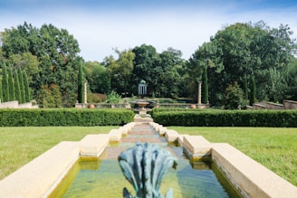 A symmetrical garden path leads to a distant structure featuring a gazebo-like design surrounded by lush greenery. The scene includes manicured hedges and a water feature in the foreground, with ornate stone pillars and pots enhancing the classical garden aesthetic.