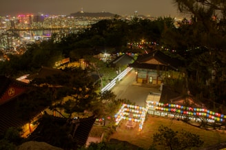 A nighttime view of a traditional Korean temple adorned with colorful lanterns set against the backdrop of a modern cityscape. The temple buildings are nestled among trees on a hill, and the bright city lights are visible in the distance.