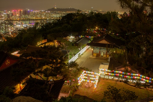 A nighttime view of a traditional Korean temple adorned with colorful lanterns set against the backdrop of a modern cityscape. The temple buildings are nestled among trees on a hill, and the bright city lights are visible in the distance.
