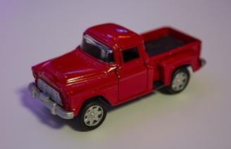 A shiny red toy pickup truck with silver accents and dark wheels is placed on a neutral surface. The truck has detailed features, including a visible grille and windows.