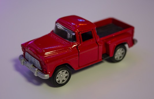A shiny red toy pickup truck with silver accents and dark wheels is placed on a neutral surface. The truck has detailed features, including a visible grille and windows.