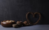 A rustic arrangement includes a variety of wooden bowls and vessels placed on a matte surface. Dried botanical elements fill the largest bowl. A heart-shaped decoration crafted from intertwined twigs is prominently displayed against a dark, textured background.