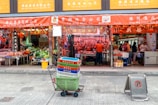 A street view of a traditional market with multiple storefronts. The stores have red signage with Chinese characters. In the center foreground, there is a cart filled with colorful crates. Behind the cart, a store displays hanging meat and other goods. People are visible inside the stores, engaged in different activities.