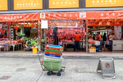 A street view of a traditional market with multiple storefronts. The stores have red signage with Chinese characters. In the center foreground, there is a cart filled with colorful crates. Behind the cart, a store displays hanging meat and other goods. People are visible inside the stores, engaged in different activities.