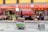 A street view of a traditional market with multiple storefronts. The stores have red signage with Chinese characters. In the center foreground, there is a cart filled with colorful crates. Behind the cart, a store displays hanging meat and other goods. People are visible inside the stores, engaged in different activities.