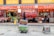 A street view of a traditional market with multiple storefronts. The stores have red signage with Chinese characters. In the center foreground, there is a cart filled with colorful crates. Behind the cart, a store displays hanging meat and other goods. People are visible inside the stores, engaged in different activities.