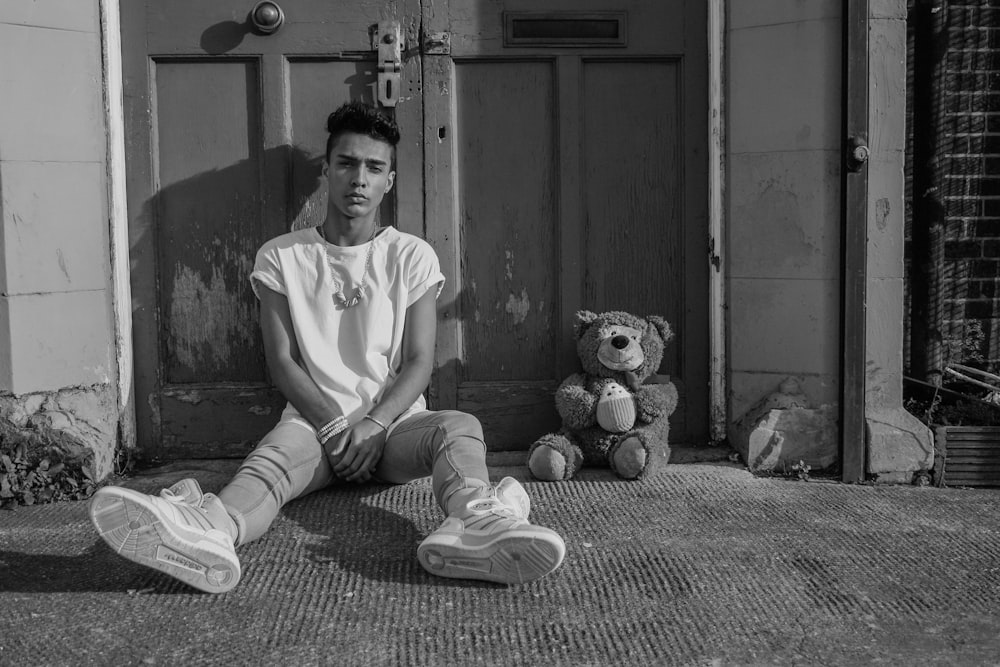 grayscale photo of boy sitting on floor beside bear plush toy