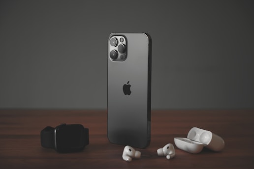 A sleek, modern smartphone with a dark metallic finish stands upright on a wooden surface. To its left lies a black smartwatch, while wireless earbuds and their open charging case are positioned to its right. The scene is set against a muted dark background, highlighting the elegance of the gadgets.
