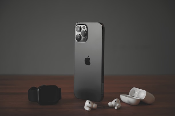 A sleek, modern smartphone with a dark metallic finish stands upright on a wooden surface. To its left lies a black smartwatch, while wireless earbuds and their open charging case are positioned to its right. The scene is set against a muted dark background, highlighting the elegance of the gadgets.